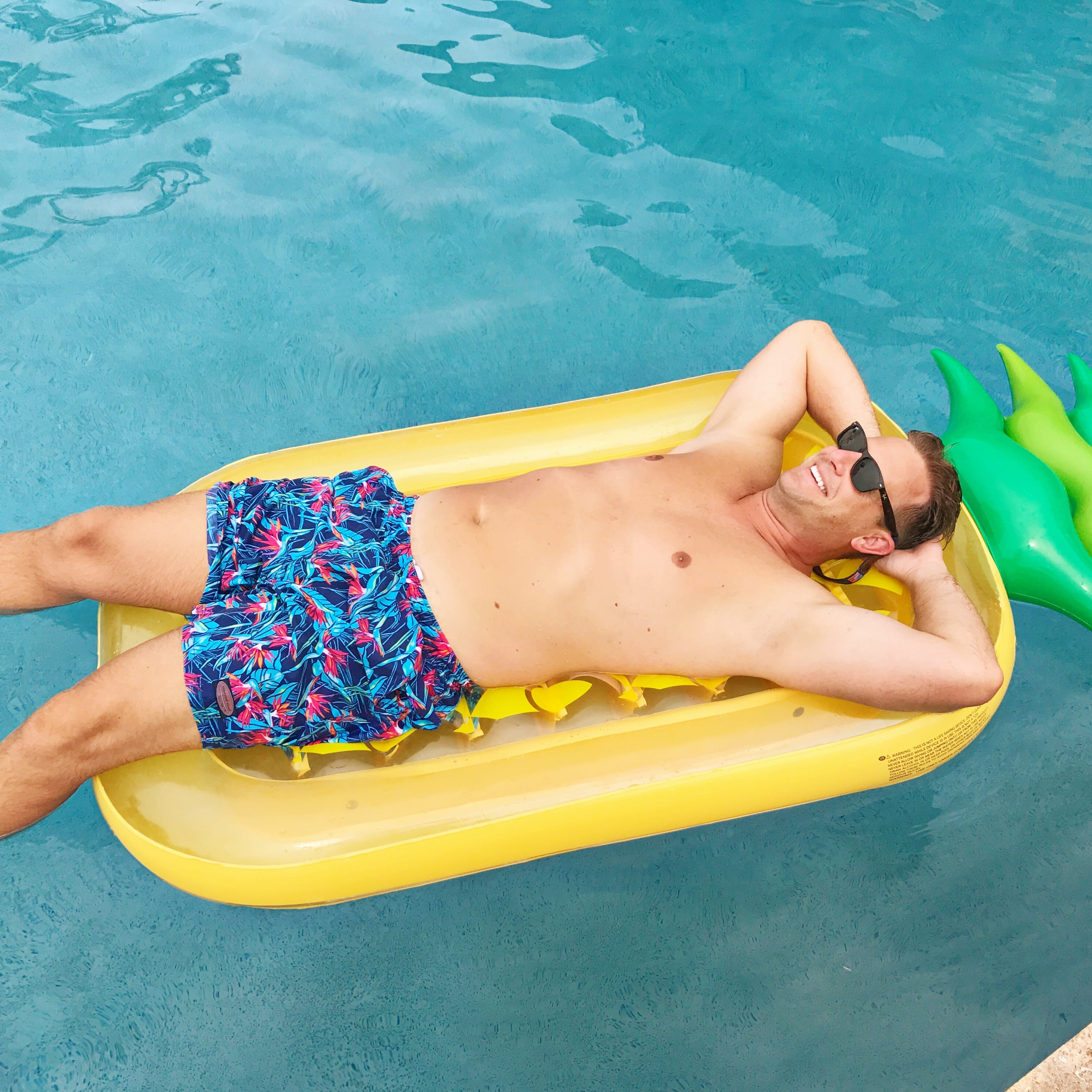Man wearing swim trunks laying on a pineapple raft.